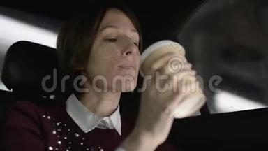 穿着白色上衣和栗色毛衣的棕色头发的漂亮女人坐在车里，喝着咖啡